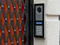 Domofon z kamerą i klawiaturą numeryczną zamontowany na ścianie obok czarnej kraty.