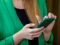 Kobieta w zielonej marynarce trzyma telefon. Szuka informacji, gdzie jest schowek w telefonie, aby ukryć ważne dane.
