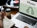 Osoba notuje pomysły na biznes, słuchając muzyki przez słuchawki. Laptop wyświetla ikonę słuchawek i napis "Music", sugerując rozpoznawanie muzyki.