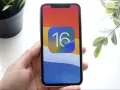 Dłoń trzyma iPhone'a z ikoną iOS 16 na kolorowej tapetcie.