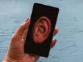 Dłoń trzyma smartfon z uchem na ekranie. Czy Twój telefon jest na podsłuchu? Sprawdź to!