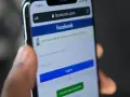Dłoń trzyma telefon z ekranem logowania Facebooka. Widoczny pasek adresu facebook.com i pola do wpisania numeru telefonu/emaila i hasła. Pomocne przy pytaniu jak odzyskać konto na FB.