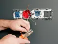 Dłonie trzymające kombinerki przy przewodach elektrycznych podłączanych do gniazdka.