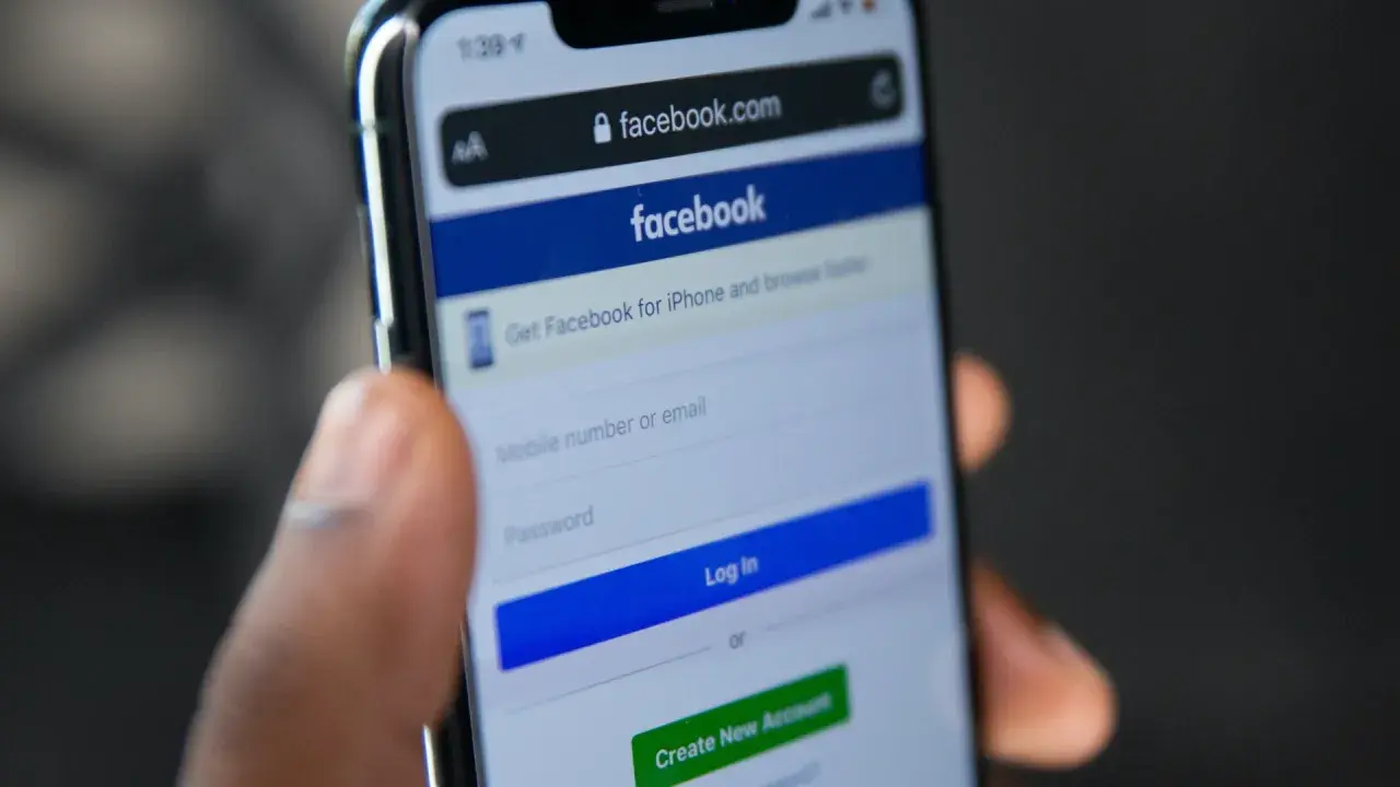 Dłoń trzyma telefon z ekranem logowania Facebooka. Widoczny pasek adresu facebook.com i pola do wpisania numeru telefonu/emaila i hasła. Pomocne przy pytaniu jak odzyskać konto na FB.