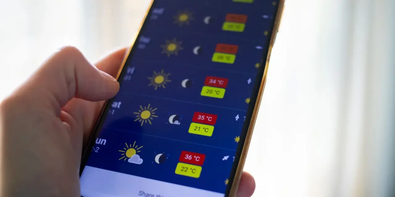 Dłoń trzyma smartfon z otwartą aplikacją pogoda, pokazującą prognozę z wysokimi temperaturami.