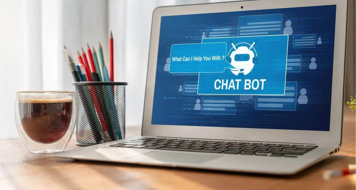 Na ekranie laptopa widać interfejs chatbota, który oferuje pomoc. Obok stoi szklanka kawy i pojemnik z przyborami do pisania.