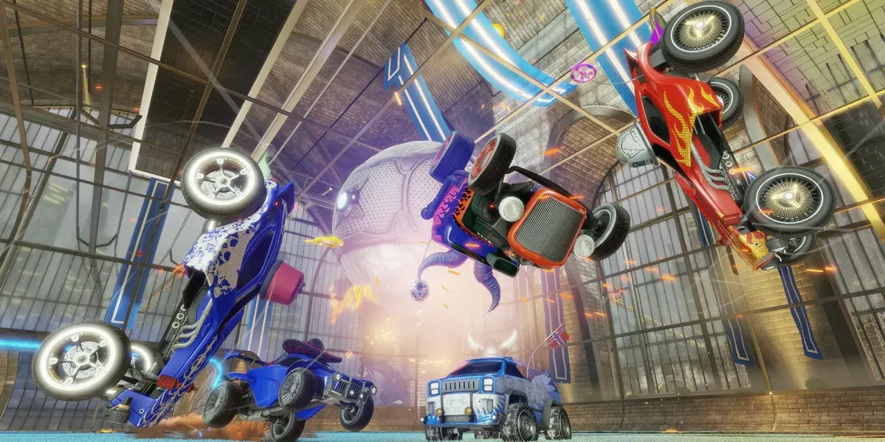 Trzy samochody z gry Rocket League w powietrzu, jeden czerwony, jeden niebieski i jeden pomarańczowy, na tle hali.