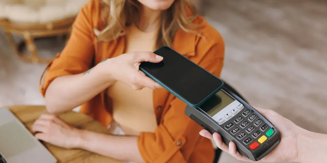 Uśmiechnięta kobieta zbliża telefon do terminala płatniczego, pokazując, jak płacić telefonem. Szybko i wygodnie.