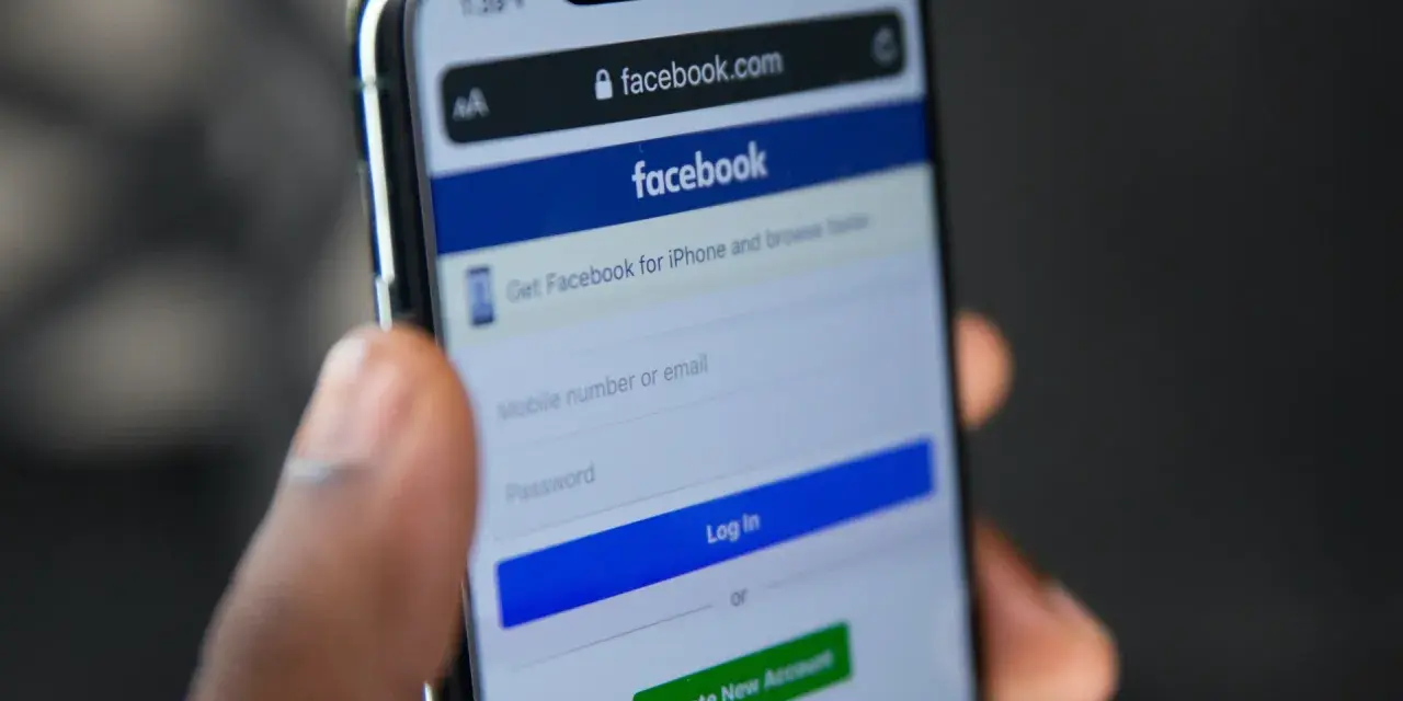 Dłoń trzyma telefon z ekranem logowania Facebooka. Widoczny pasek adresu facebook.com i pola do wpisania numeru telefonu/emaila i hasła. Pomocne przy pytaniu jak odzyskać konto na FB.