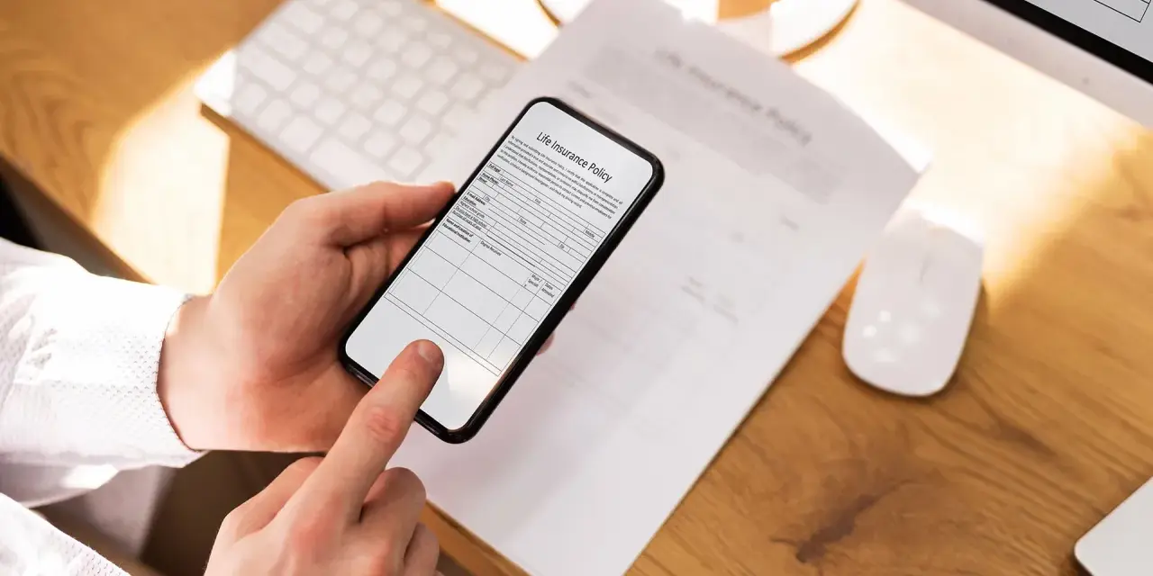 Ręce trzymają smartfon, który działa jak skaner dokumentów, skanując polisę ubezpieczeniową leżącą na biurku.