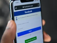 Dłoń trzyma telefon z ekranem logowania Facebooka. Widoczny pasek adresu facebook.com i pola do wpisania numeru telefonu/emaila i hasła. Pomocne przy pytaniu jak odzyskać konto na FB.