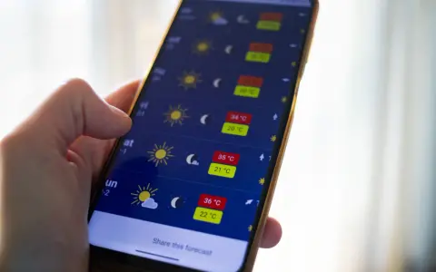 Dłoń trzyma smartfon z otwartą aplikacją pogoda, pokazującą prognozę z wysokimi temperaturami.