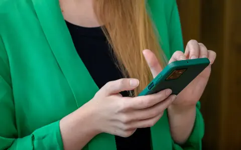 Kobieta w zielonej marynarce trzyma telefon. Szuka informacji, gdzie jest schowek w telefonie, aby ukryć ważne dane.