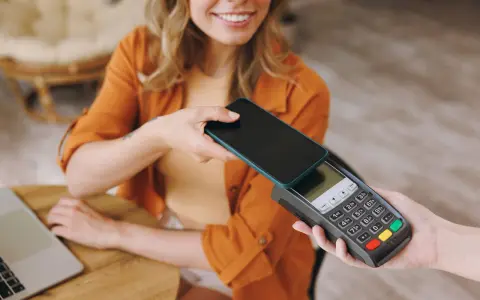 Uśmiechnięta kobieta zbliża telefon do terminala płatniczego, pokazując, jak płacić telefonem. Szybko i wygodnie.
