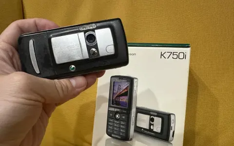 Dłoń trzyma czarny telefon Sony Ericsson K750i, obok stoi pudełko z tym modelem.
