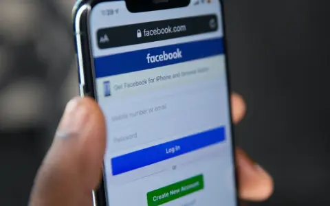 Dłoń trzyma telefon z ekranem logowania Facebooka. Widoczny pasek adresu facebook.com i pola do wpisania numeru telefonu/emaila i hasła. Pomocne przy pytaniu jak odzyskać konto na FB.