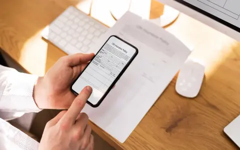 Ręce trzymają smartfon, który działa jak skaner dokumentów, skanując polisę ubezpieczeniową leżącą na biurku.