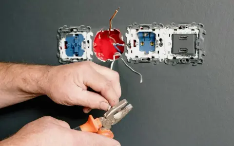 Dłonie trzymające kombinerki przy przewodach elektrycznych podłączanych do gniazdka.