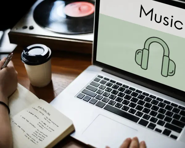 Osoba notuje pomysły na biznes, słuchając muzyki przez słuchawki. Laptop wyświetla ikonę słuchawek i napis "Music", sugerując rozpoznawanie muzyki.