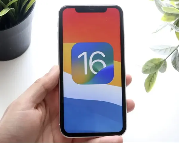 Dłoń trzyma iPhone'a z ikoną iOS 16 na kolorowej tapetcie.
