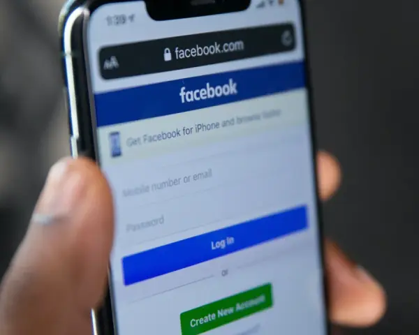 Dłoń trzyma telefon z ekranem logowania Facebooka. Widoczny pasek adresu facebook.com i pola do wpisania numeru telefonu/emaila i hasła. Pomocne przy pytaniu jak odzyskać konto na FB.