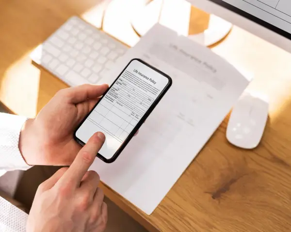 Ręce trzymają smartfon, który działa jak skaner dokumentów, skanując polisę ubezpieczeniową leżącą na biurku.