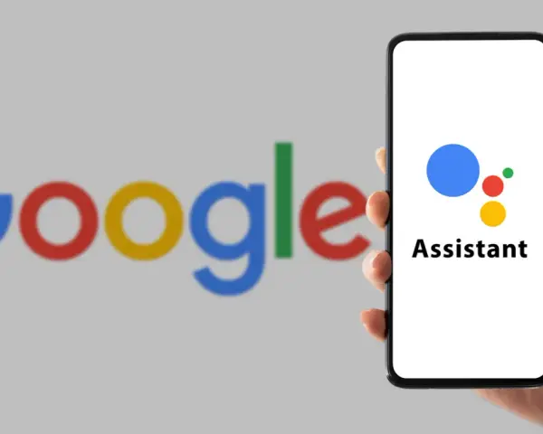 Dłoń trzyma smartfon z logo Asystenta Google na ekranie, w tle logo Google.