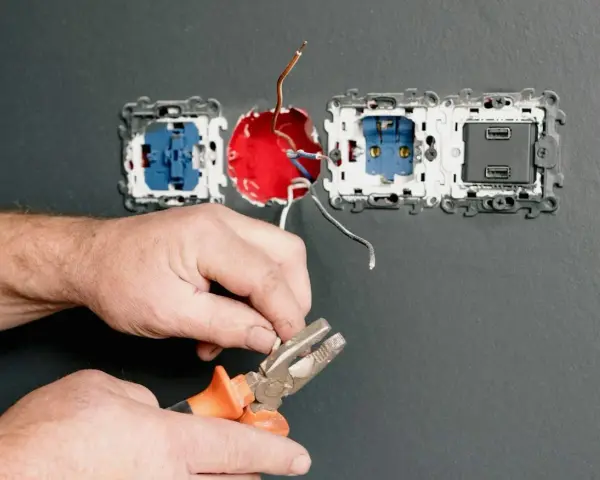 Dłonie trzymające kombinerki przy przewodach elektrycznych podłączanych do gniazdka.