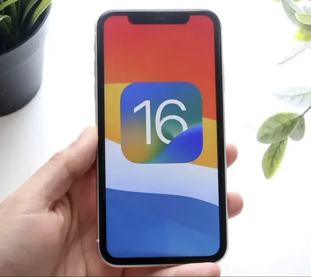 Dłoń trzyma iPhone'a z ikoną iOS 16 na kolorowej tapetcie.