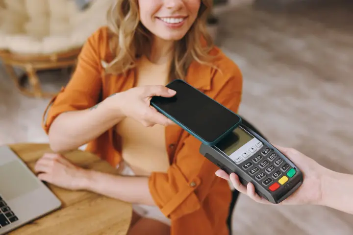 Uśmiechnięta kobieta zbliża telefon do terminala płatniczego, pokazując, jak płacić telefonem. Szybko i wygodnie.