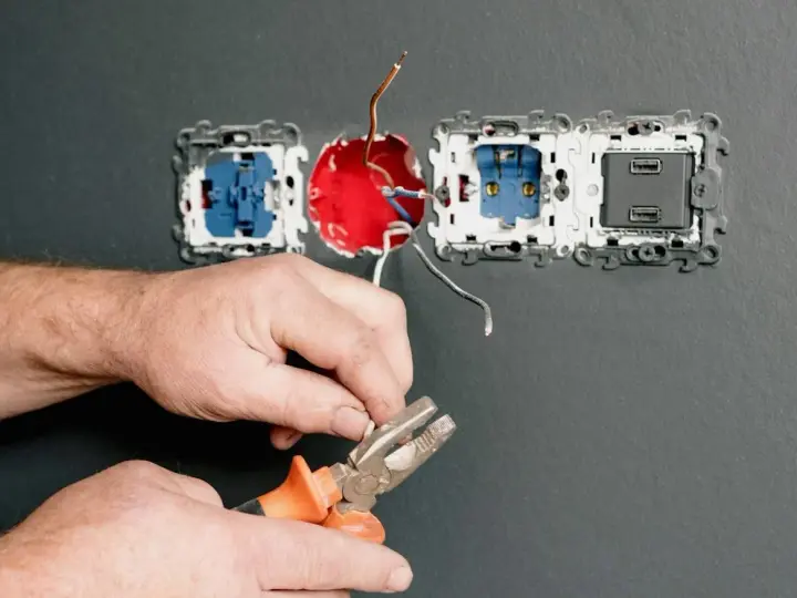 Dłonie trzymające kombinerki przy przewodach elektrycznych podłączanych do gniazdka.