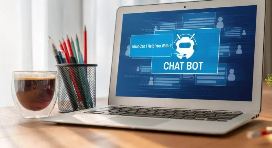 Na ekranie laptopa widać interfejs chatbota, który oferuje pomoc. Obok stoi szklanka kawy i pojemnik z przyborami do pisania.