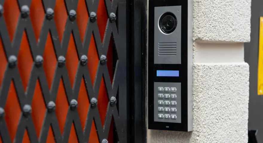 Domofon z kamerą i klawiaturą numeryczną zamontowany na ścianie obok czarnej kraty.