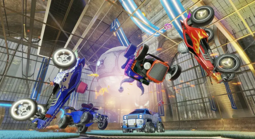 Trzy samochody z gry Rocket League w powietrzu, jeden czerwony, jeden niebieski i jeden pomarańczowy, na tle hali.
