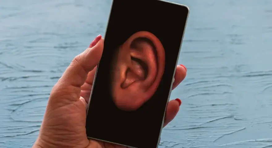 Dłoń trzyma smartfon z uchem na ekranie. Czy Twój telefon jest na podsłuchu? Sprawdź to!
