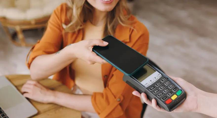 Uśmiechnięta kobieta zbliża telefon do terminala płatniczego, pokazując, jak płacić telefonem. Szybko i wygodnie.