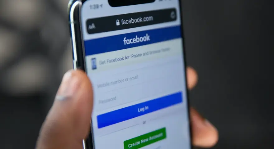 Dłoń trzyma telefon z ekranem logowania Facebooka. Widoczny pasek adresu facebook.com i pola do wpisania numeru telefonu/emaila i hasła. Pomocne przy pytaniu jak odzyskać konto na FB.