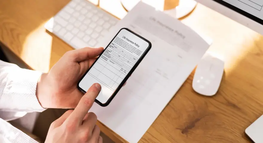 Ręce trzymają smartfon, który działa jak skaner dokumentów, skanując polisę ubezpieczeniową leżącą na biurku.