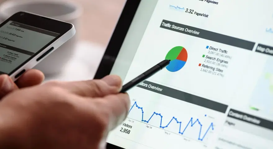 Dłoń z rysikiem wskazuje na wykres kołowy i liniowy na ekranie tabletu, obok smartfona.